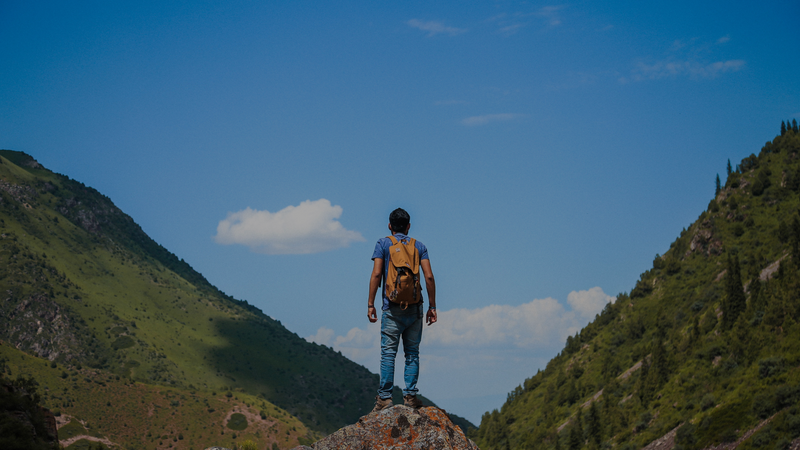 Person with a backpack standing on a mountain peak with a clear blue sky and green mountains in the background.