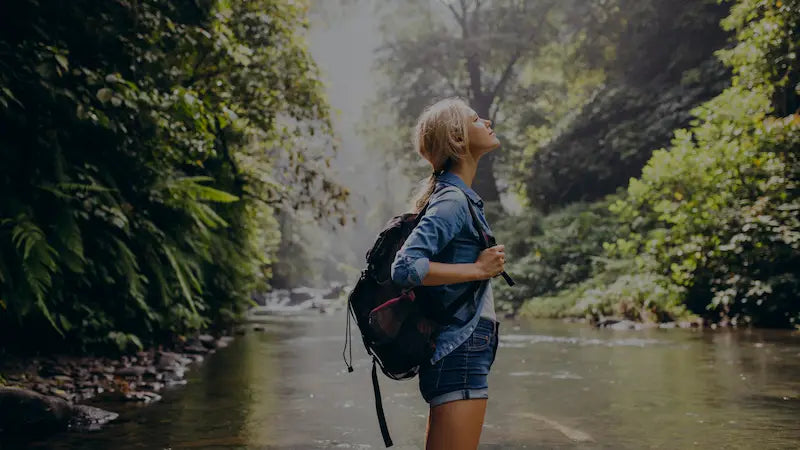 Woman with a backpack standing by a river surrounded by greenery