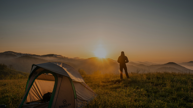 Person standing on a hill with a tent at sunrise or sunset, surrounded by mountains.